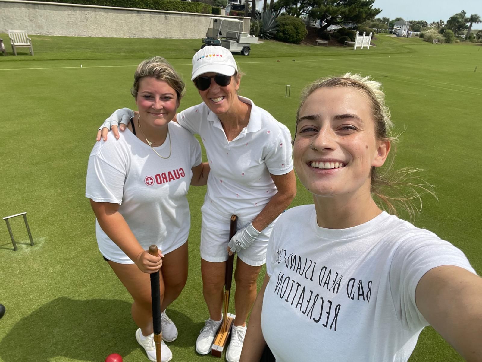 Three smiling women pose on a croquet lawn, holding mallets.