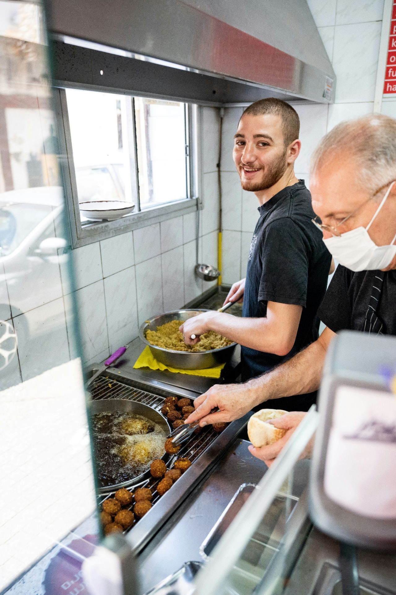 A smiling young man in a black shirt cooks food behind a counter while an older man wearing a mask looks on.