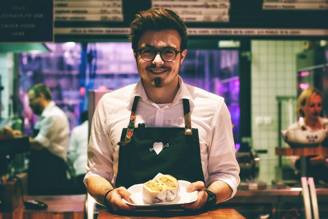 Man wearing glasses and an apron holds a plate with a pastry.