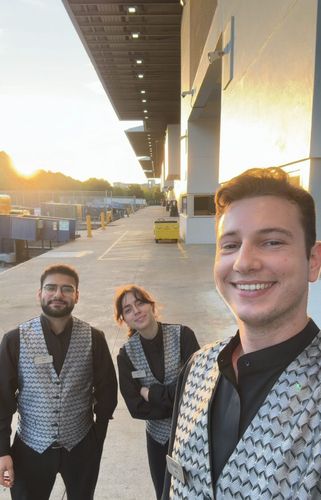 Three men in formal vests and shirts pose for a selfie outdoors at sunset.