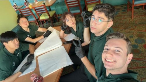 A group of five young adults in green shirts and black gloves are gathered around a table.