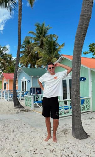 Man in white t-shirt and black shorts leans against a palm tree on a sandy beach near colorful beach bungalows.