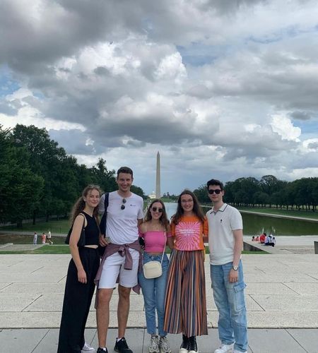 Five young adults pose for a photo with the Washington Monument in the background on a cloudy day.