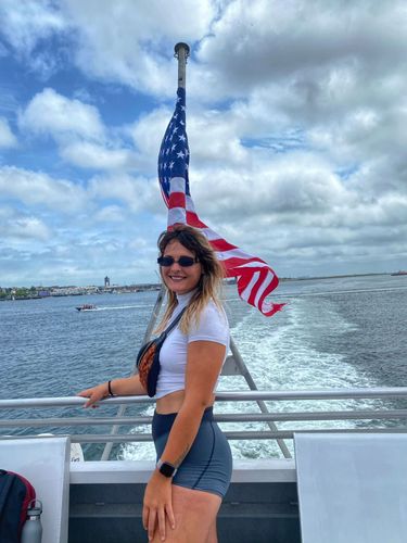 Woman in sunglasses, white top, and denim shorts stands on a boat deck with an American flag flying behind her.