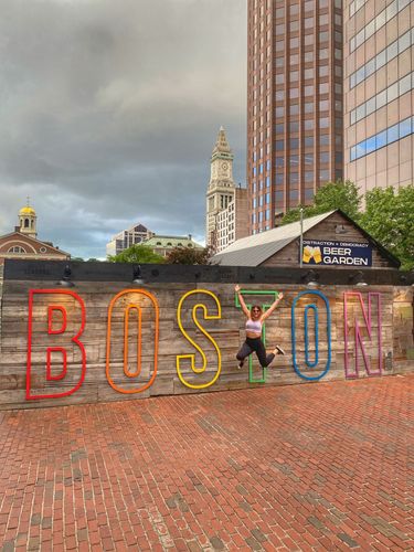 Person jumps through the letters of a large "BOSTON" sign with brickwork and city buildings in the background.