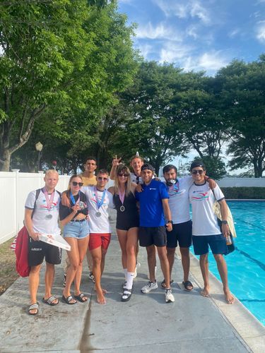 A group of nine lifeguards in swim attire poses for a photo by a swimming pool on a sunny day.