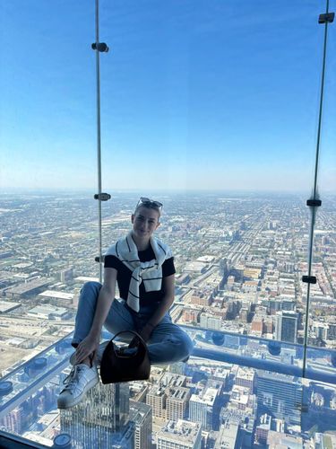 A person sits on a glass floor overlooking a cityscape from a tall building.