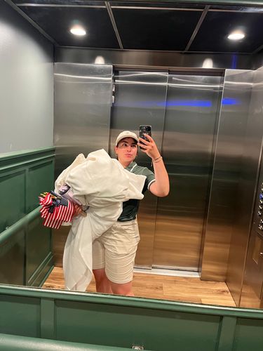 Person in a baseball cap takes an elevator selfie, holding a large white bag.
