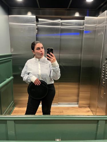 Woman in white shirt and dark pants taking a mirror selfie in an elevator.