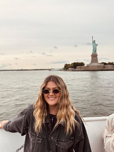 Young woman with long blonde hair and sunglasses smiles on a boat with the Statue of Liberty in the background.